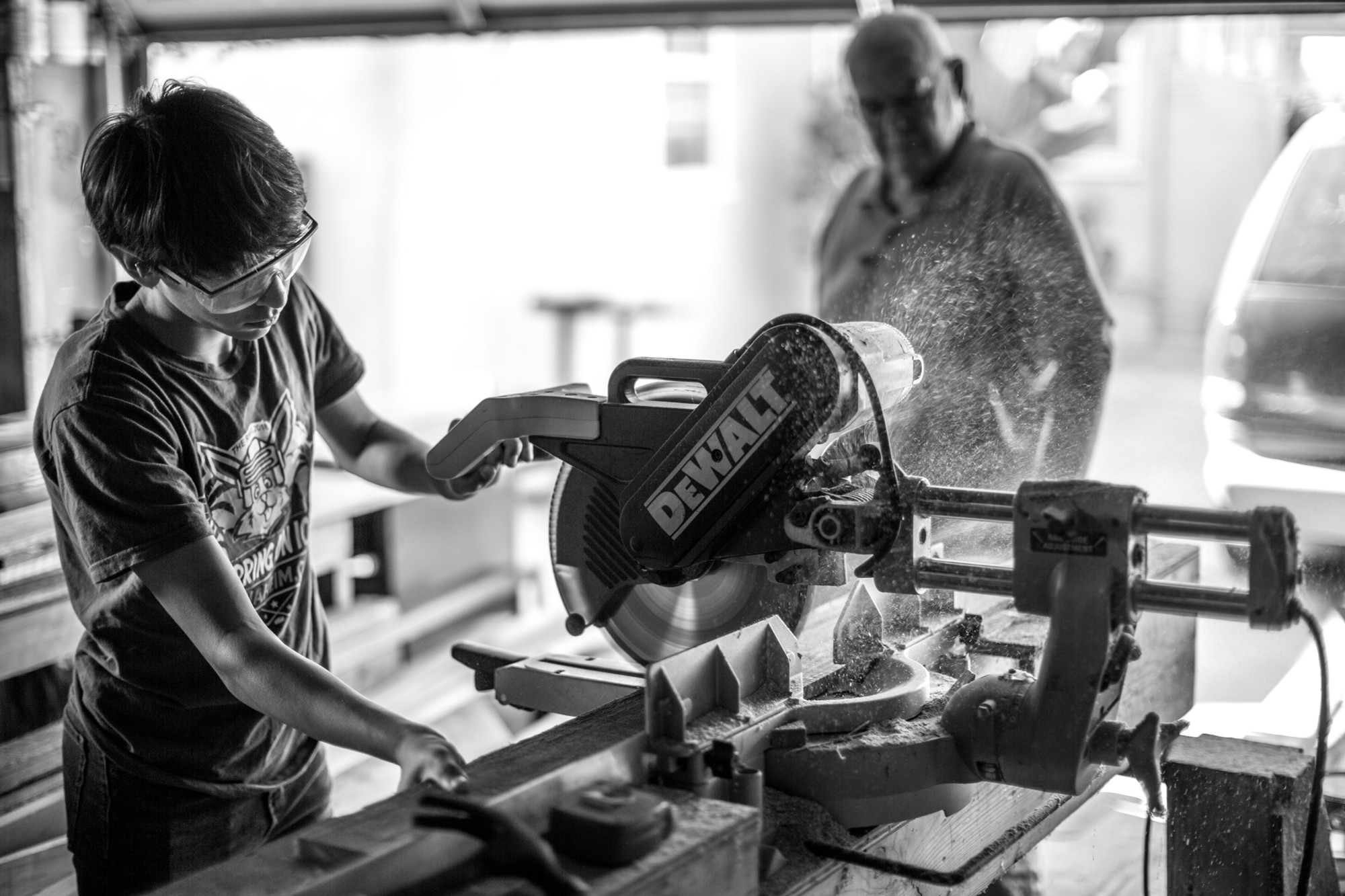A young man in protective goggles, observed by a tutor, cuts come wood with an industrial, electric saw