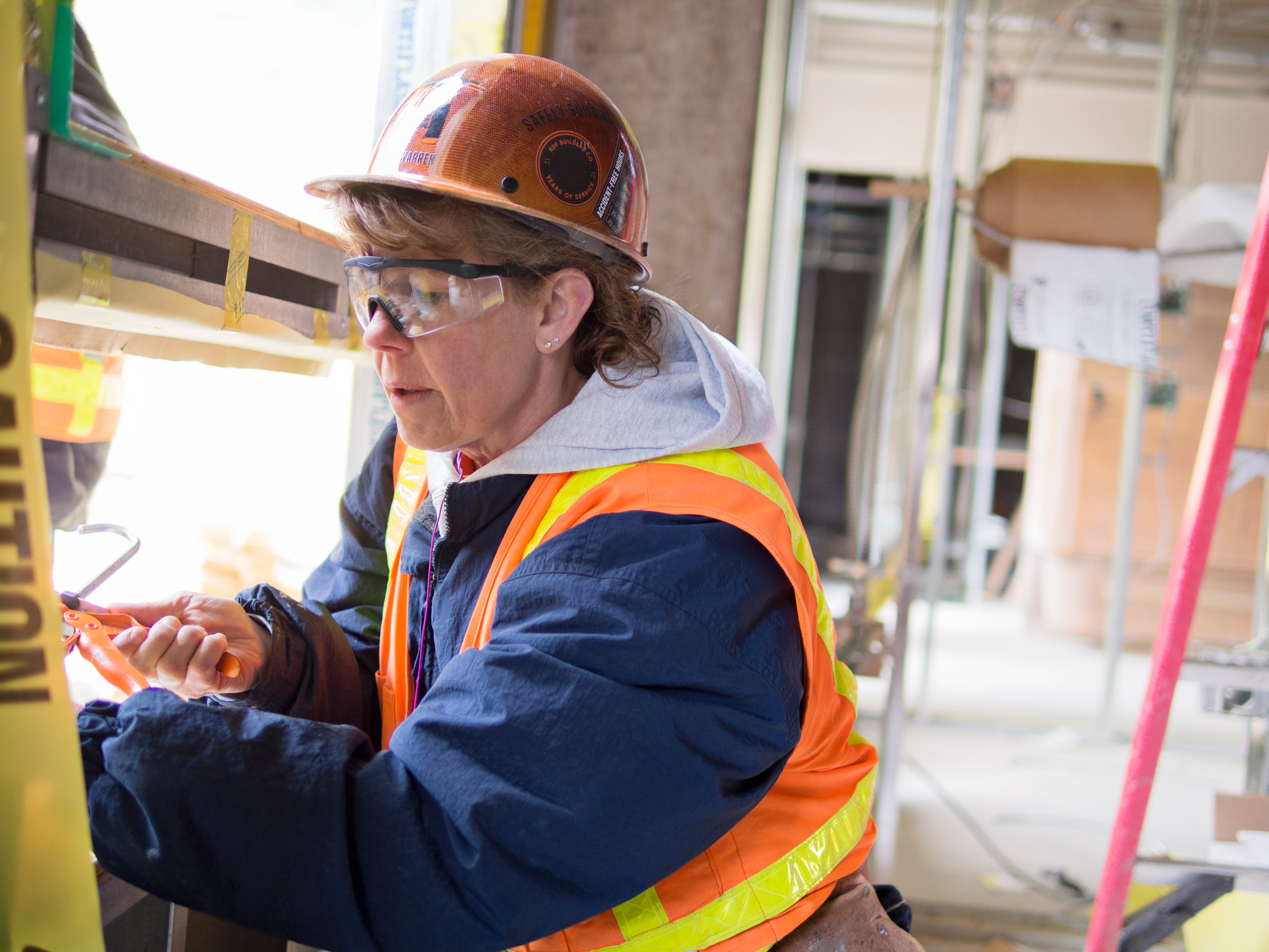 An older woman wears a hard hat and fluorescent jacket, and protective glasses. She works in a building site.