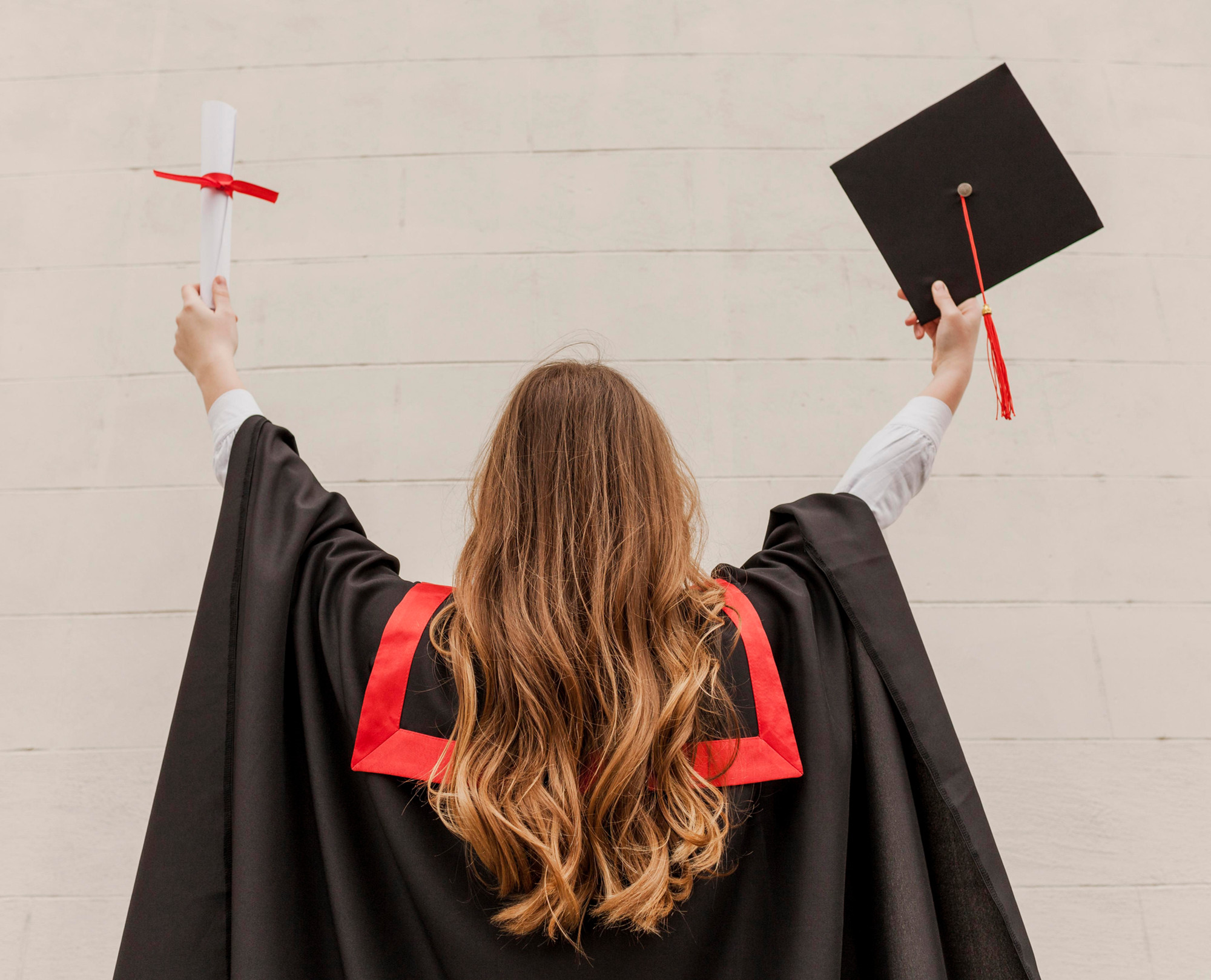The back of a woman in a graduating gown holding a graduate cap in her right hand above her head and her scrolled-up degree in her left above her head.