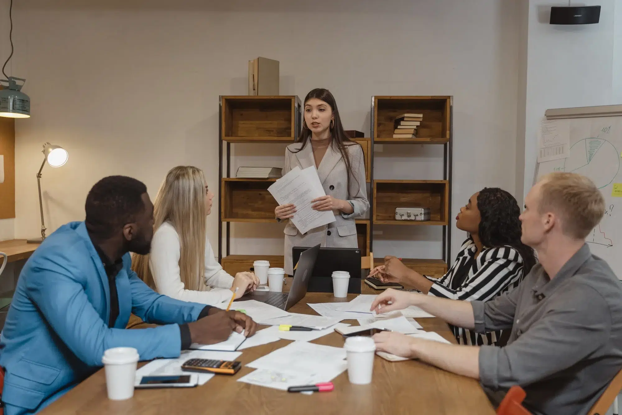 A woman in a suit addresses her team in a business meeting. The table is adorned with papers, notebooks, coffee cups and devices. They all listen to her intently.