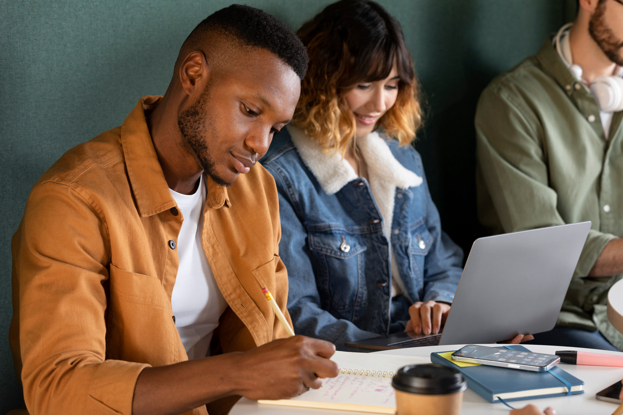 Three young people work side by side in a cafe. Their notebooks, devices, coffee cups and highlighters sit on the round table in front of them.