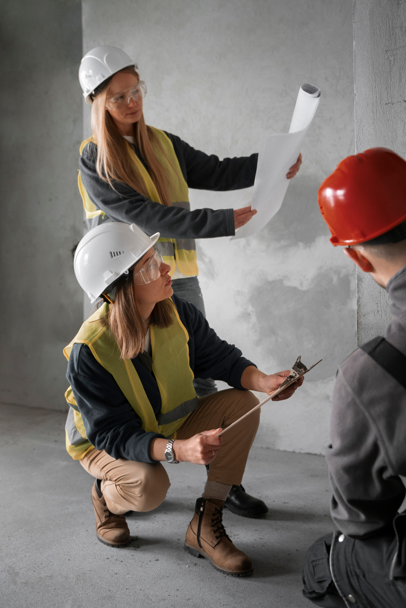 Two women examine a wall, wearing hard hats and fluorescent jackets in a construction site.
