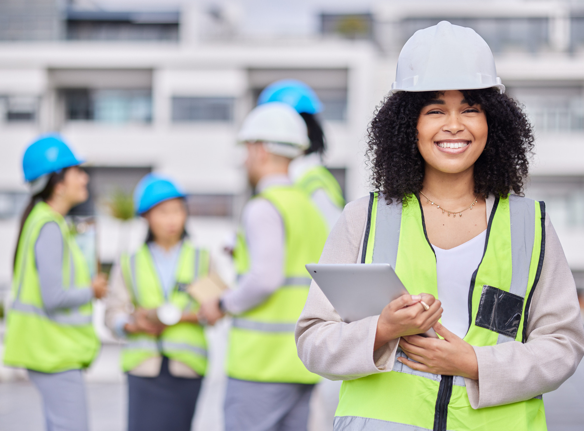 A woman working in construction smiles wearing a hard hat and fluorescent jacket cradling a tablet device. Behind her are 4 of her colleagues.