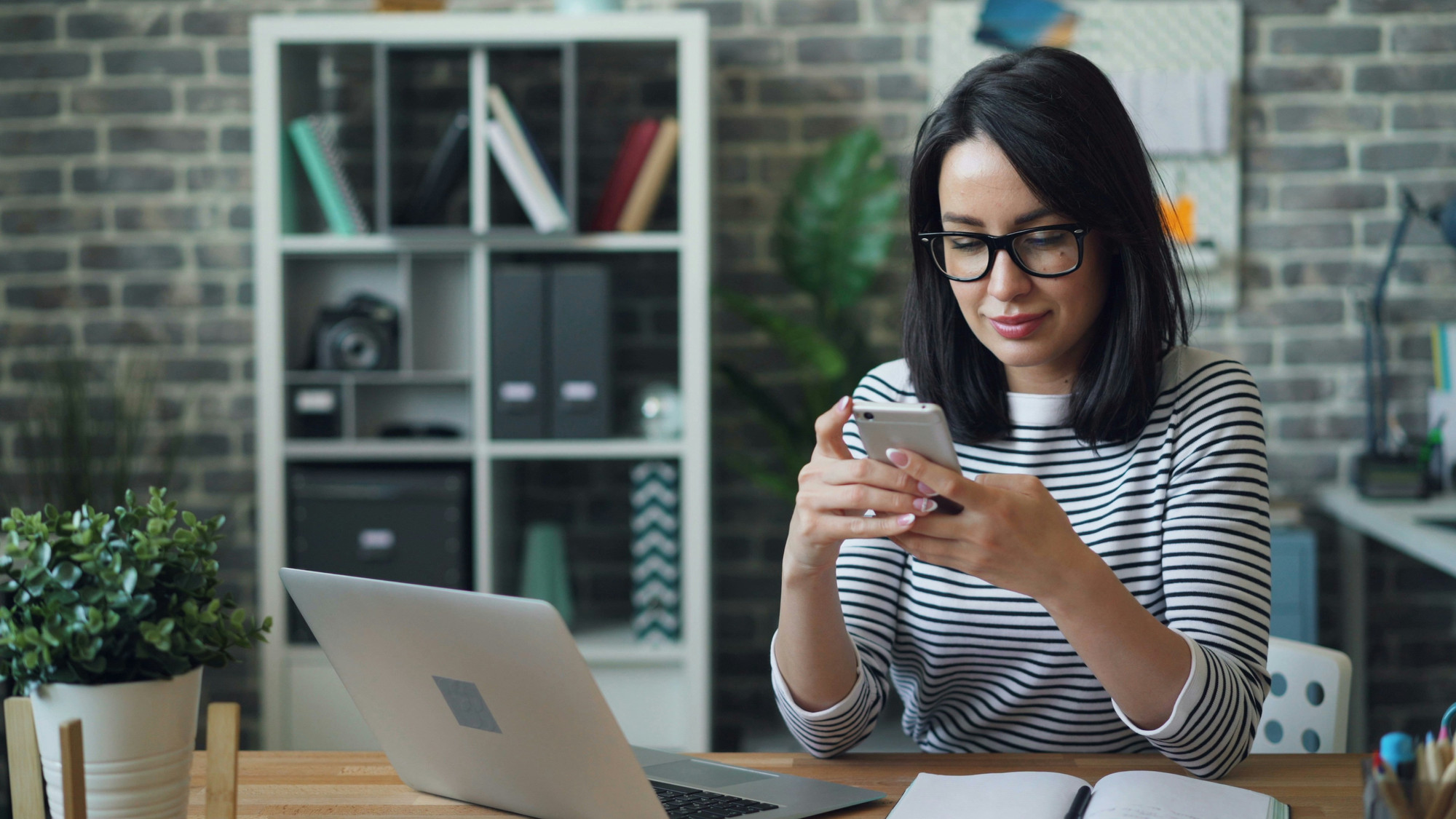 A woman texts on her smartphone next to her open laptop in an office with an exposed brick wall.