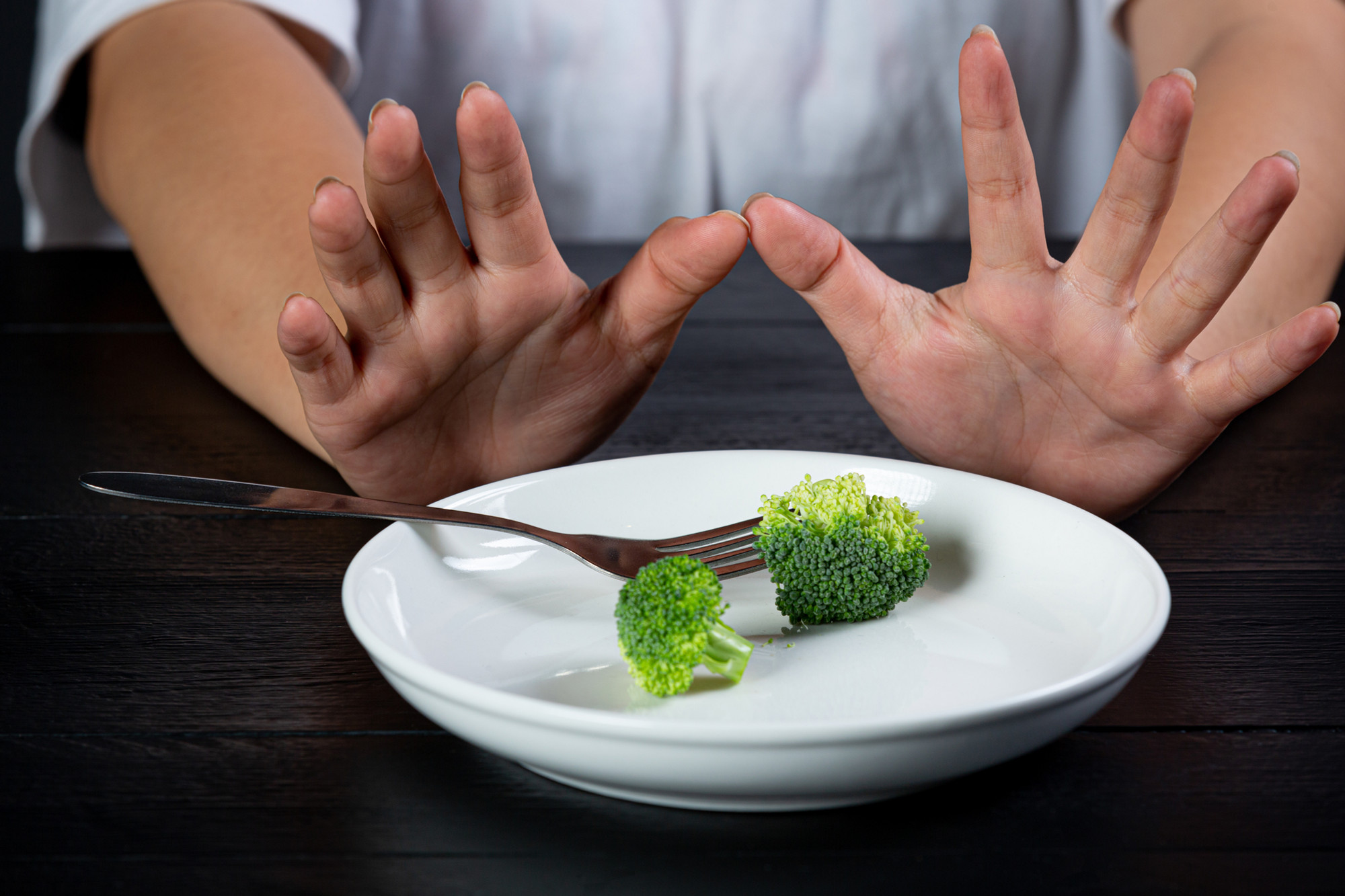 A pair of hands push a way a plate of two broccolli piece on a white plate