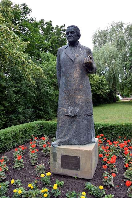 A large statue of a mature woman making a gentle fist with her left hand. The statue sits in a bed of flowers in a green park.