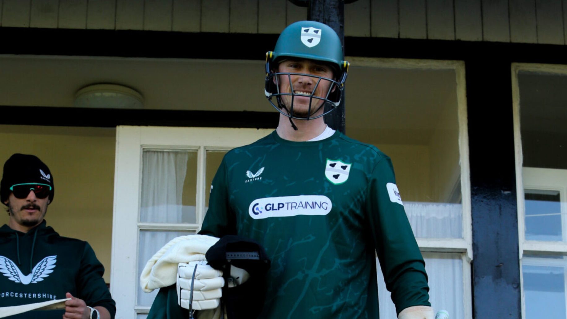 A cricket player of Worcestershire County Cricket Club wears a GLP Training branded shirt and smiles wearing protective head wear.