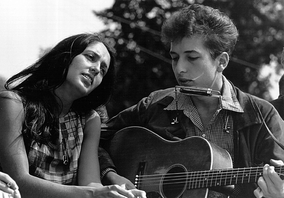 A man wearing a harmonica and playing the guitar. A woman sits by him, leaning her body toward him whilst she sings. She wears a 'peace; symbol chain around her neck.