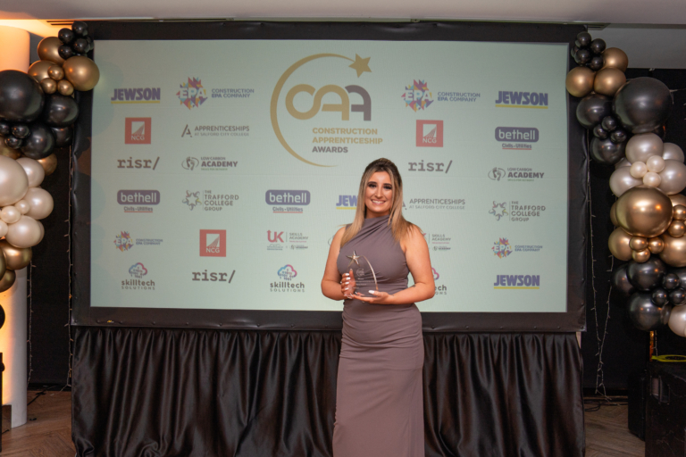 Alyssa Howard - a white woman with long, dark blonde hair - poses with an award in formal wear in front of a display of industry logos