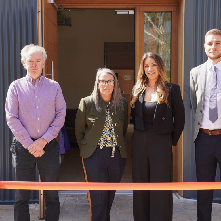 4 of our team stand in front of the building waiting to cut an orange ribbon and declare it open.