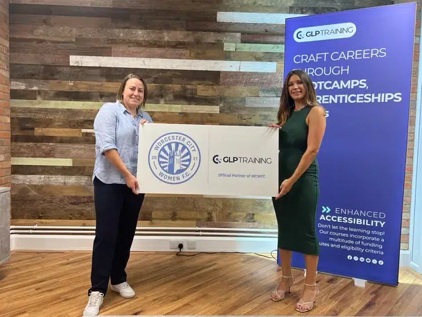 Two women in formal wear hold a large rectangular sign together that reads 'Worcester City Women FC x GLP Training Official Partner of WCWFC"