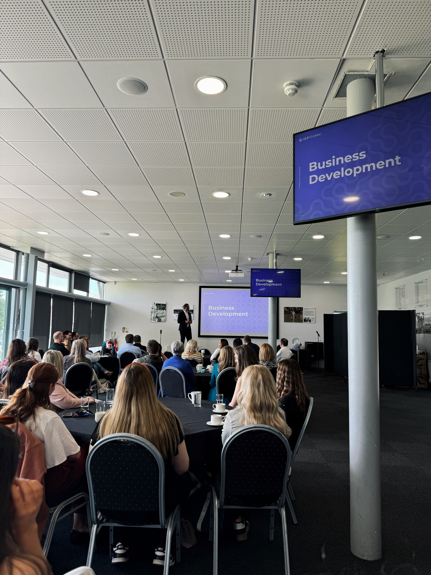 The Business Development section of the GLP Training annual conference in a conference room. A man in a suit looks toward the screen whilst addressing the room from the stage.