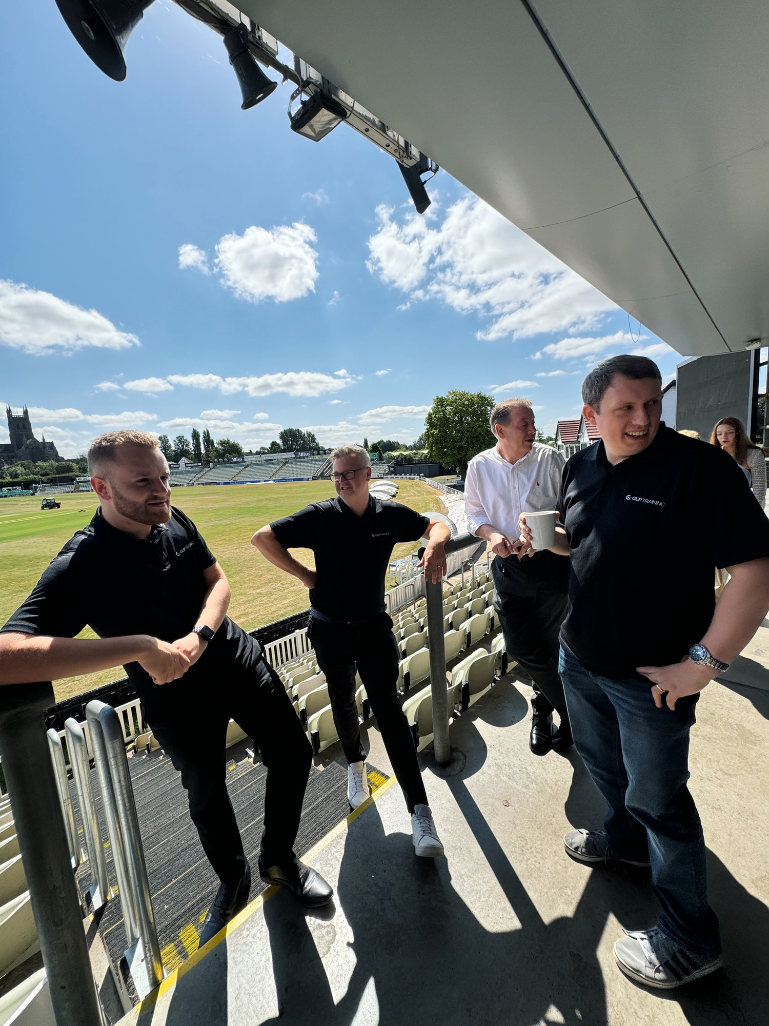 A group of 4 men lean on the metal bar of the cricket stadium's balcony on a sunny afternoon. One drinks a coffee.
