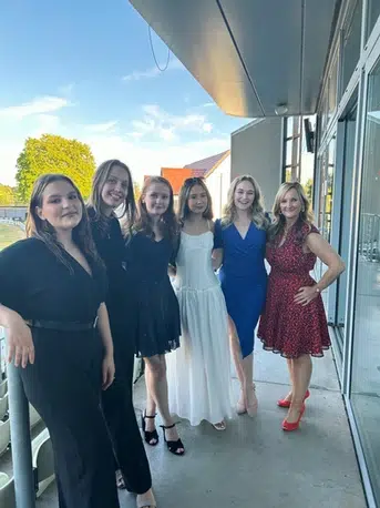 A group of 6 women dressed in formal wear on a balcony posing for the camera