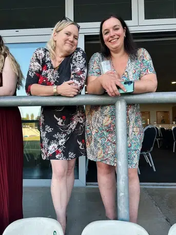 Two women in formal wear lean on a metal bar smiling at the camera