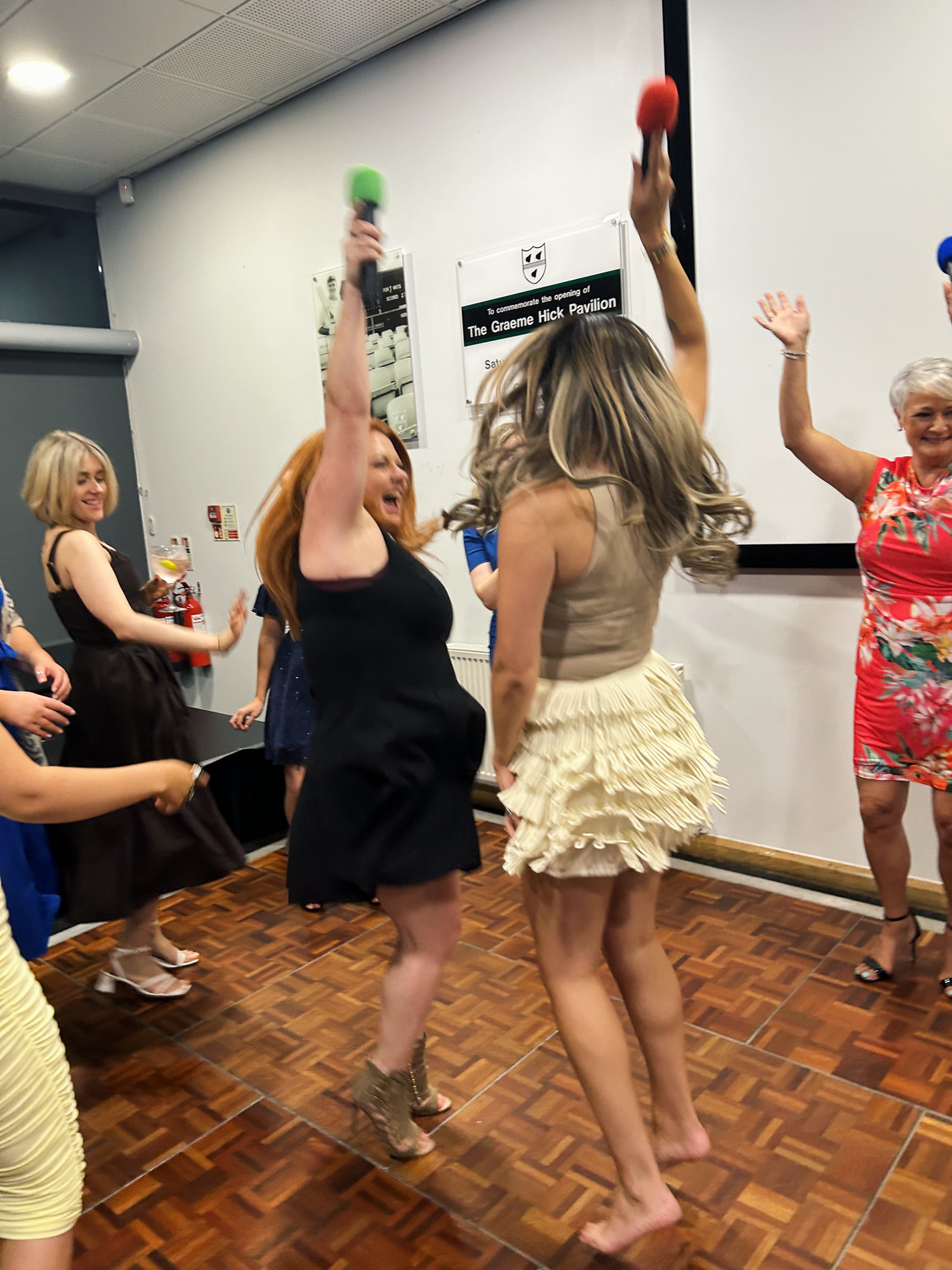 A group of female colleagues in formal wear dance together on a dance floor. One has removed her shoes.