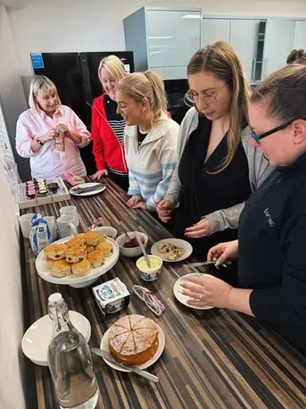 A group of 5 women gather around a station in a shared kitchen to help themselves to cake and scones and tea.