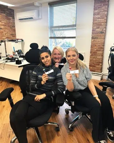 A group of three woman in office chairs pose for the camera with white slips of paper and a lolly pop each
