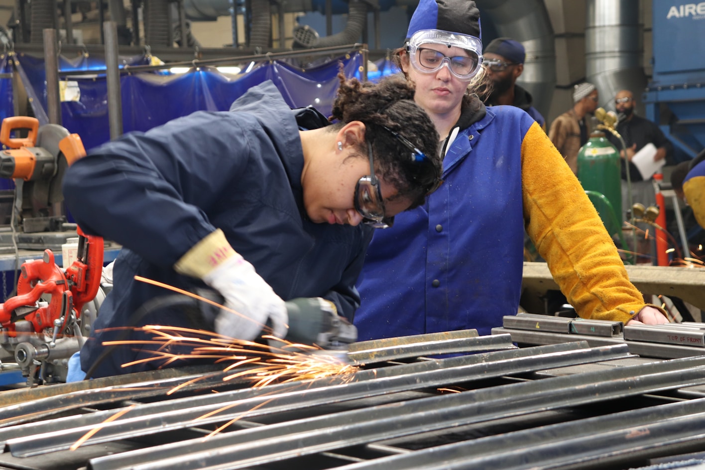 Two female students in protective clothes and goggles perform metal work; sparks fly from the metal grinder meeting the worked on metal.