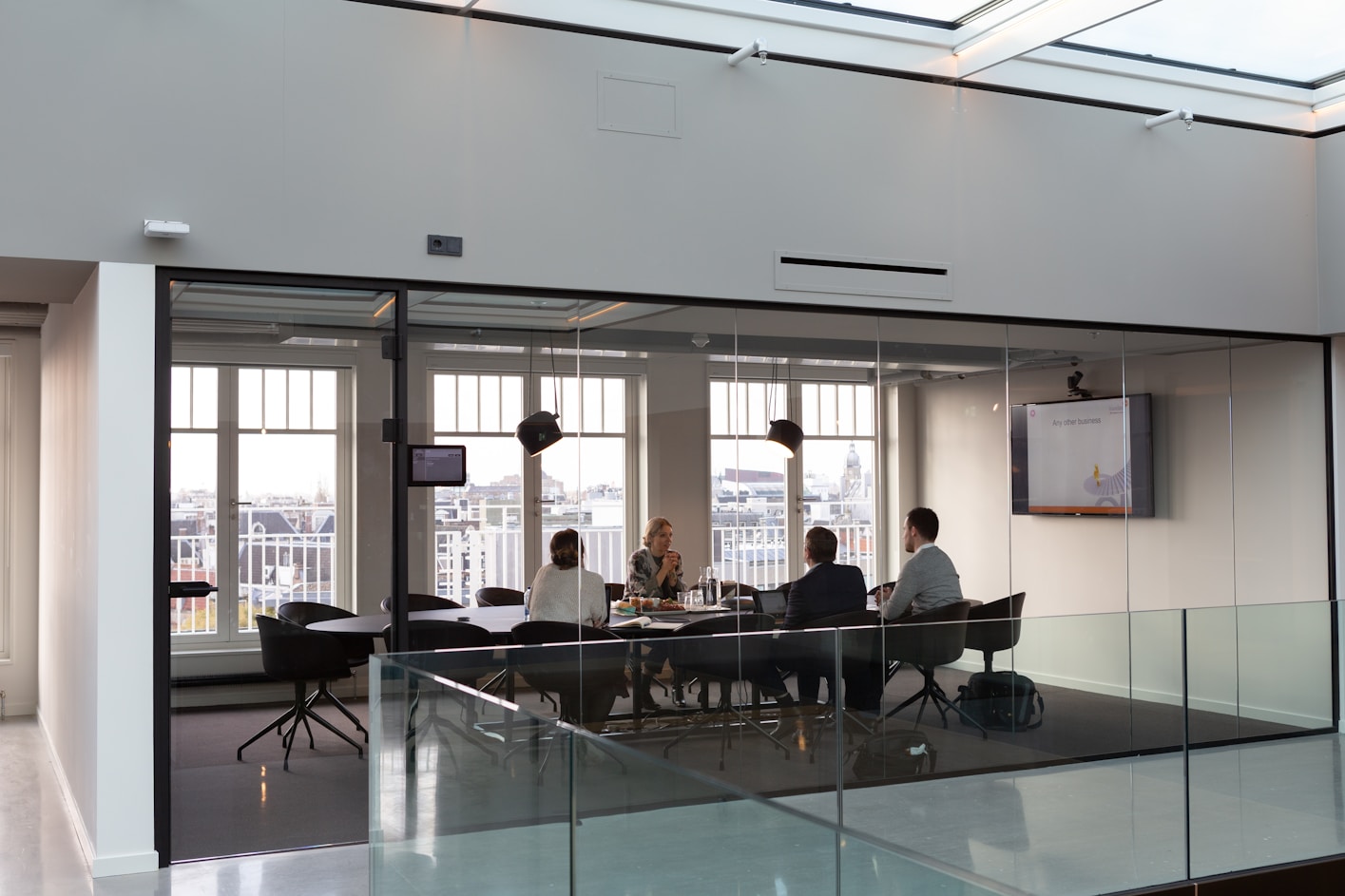 A wide shot of a meeting taking place between 4 colleagues in business wear around a large conference table in well-lit, open plan office room with glass walls and large windows