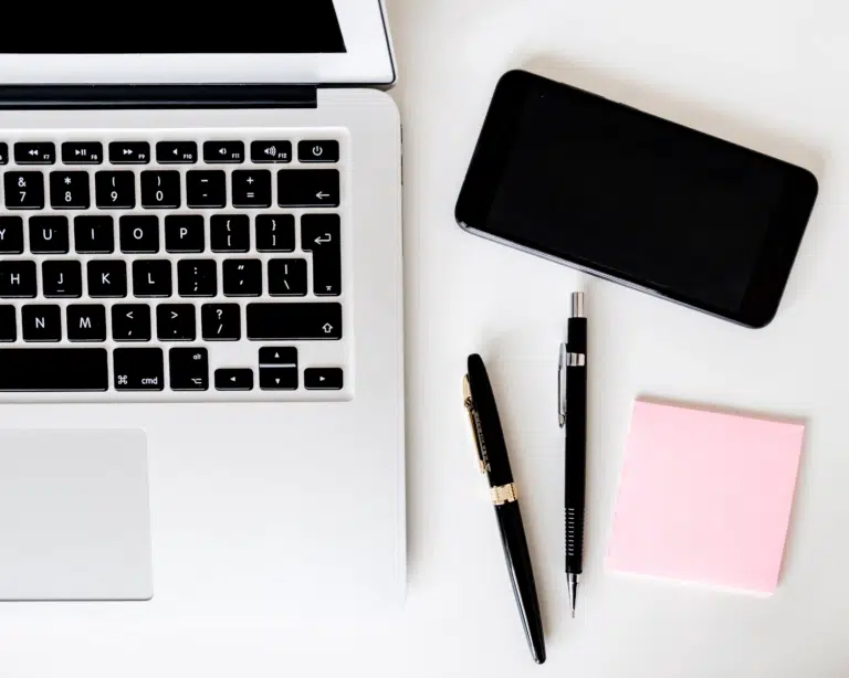 A birds eye view, close, of a laptop, smartphone, two pens and a pink post it note.