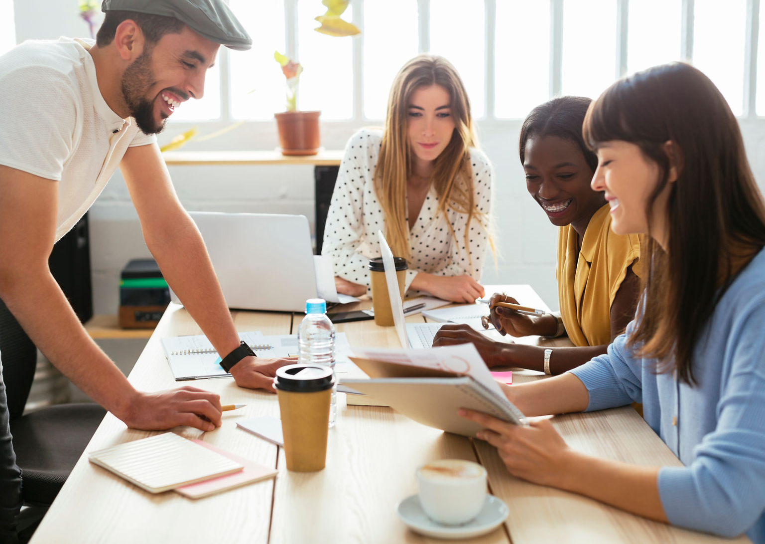 A diverse group of 4 young people work collaboratively over a wooden table adorned with coffee cups, mugs, papers and a laptop. They all smile