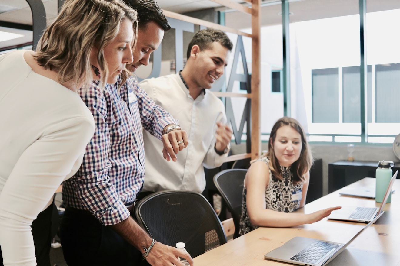 A diverse group of white colleagues gather around a laptop and have a discussion in a well-lit office