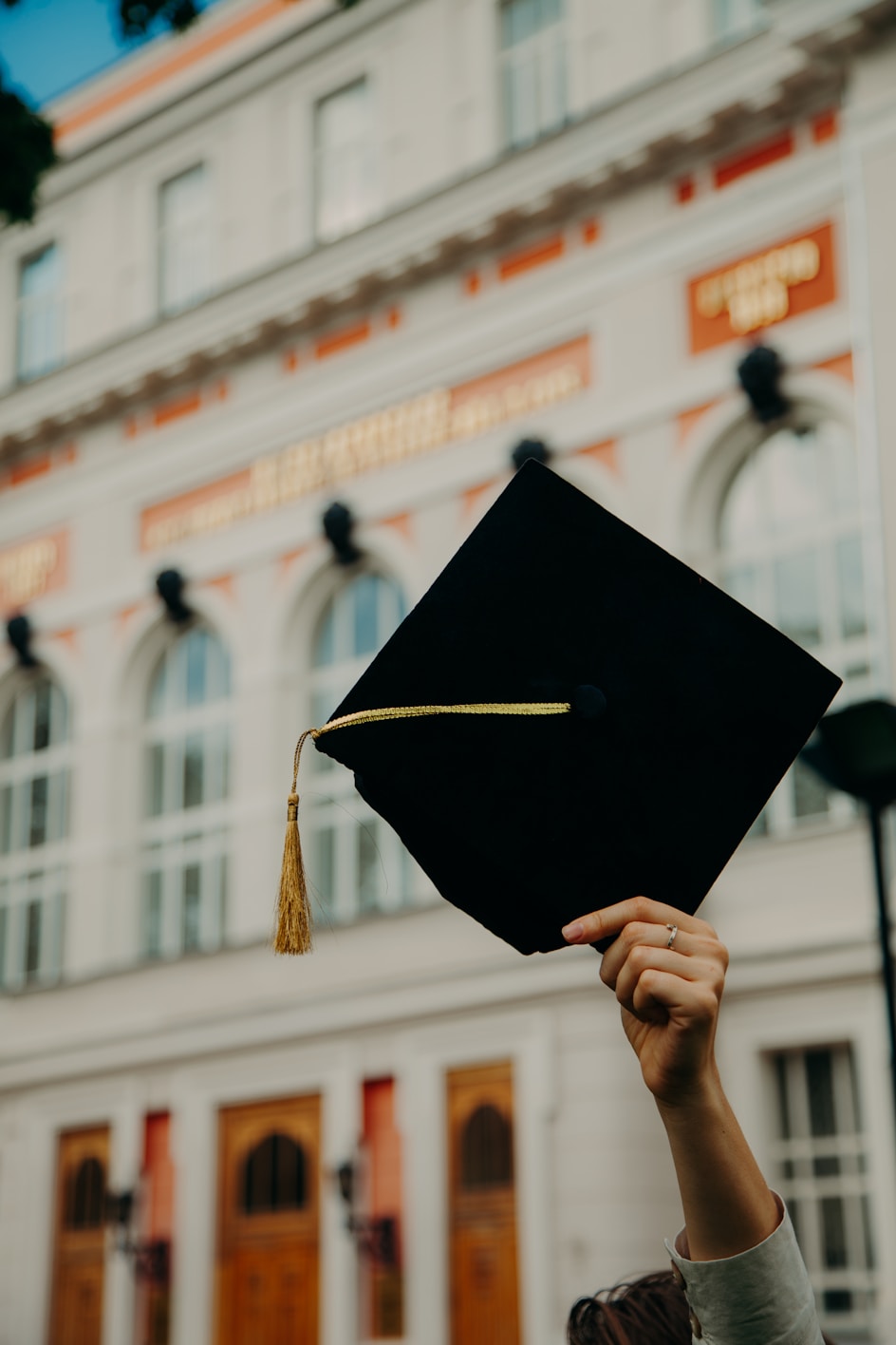 A close up of a black graduation hat being held high in front of a grand looking building. The hat has a gold/yellow tassle.