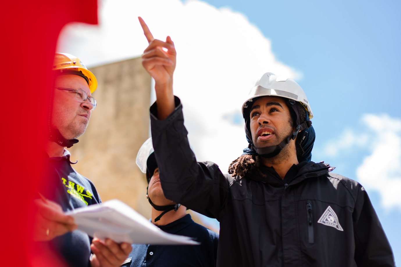 A young black male points upward wearing a black hoodie and a white hard hat. A white, maturer male listens to him whilst holding a piece of paper and also wearing a hard hat. They are outside.
