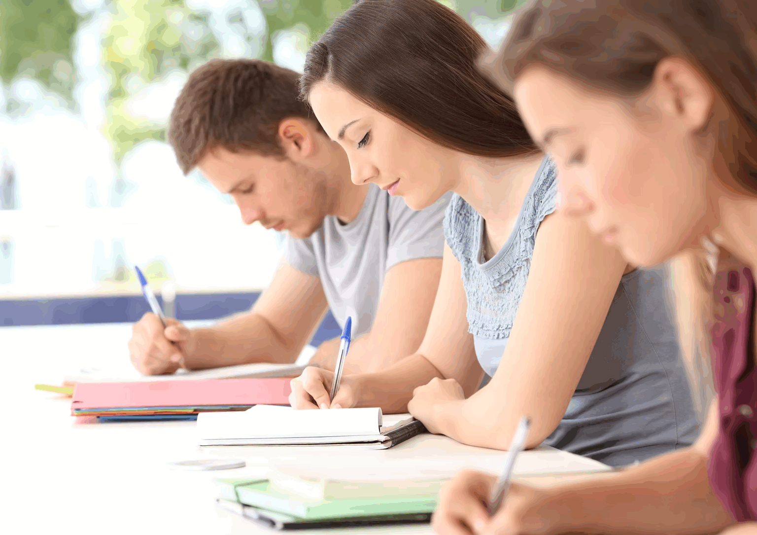 Three white students, one male and two female, make notes with pen and paper. They all are engrossed in their work