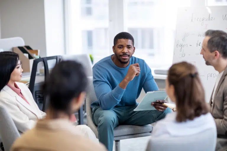 A black, male in a blue jumper refers to a clipboard in his hands whilst talking to 4 other people in a circle. Behind him is a white board that indicates 'Irregular Verbs'