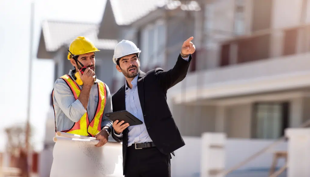 A manager of a construction site points out in a collaborative manor with his site working colleague who speaks into a walkie talkie.