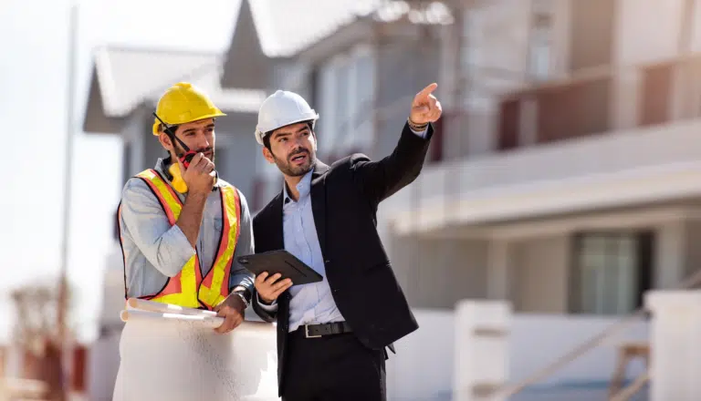 A manager of a construction site points out in a collaborative manor with his site working colleague who speaks into a walkie talkie. Both wear shirts and hard hats.