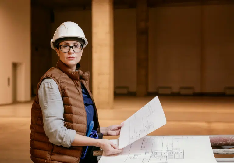 A female construction manager wearing a gilet and hard hat and glasses looks directly at camera whilst holding floorplans. She's in a large construction room.