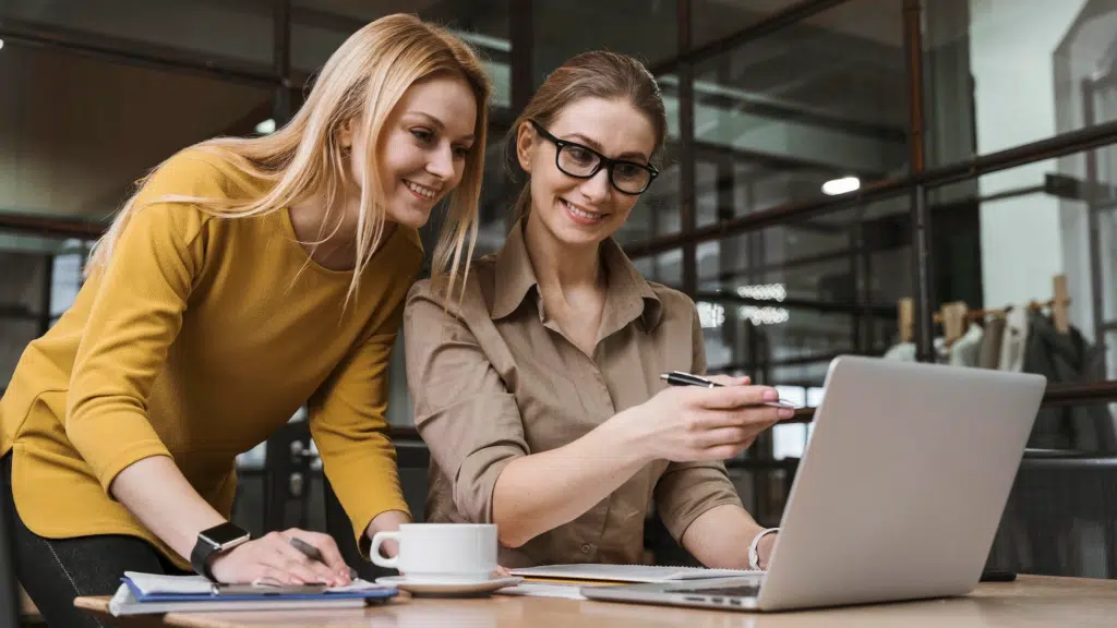 Two female works collaborate over a laptop in an office environment. They both smile.