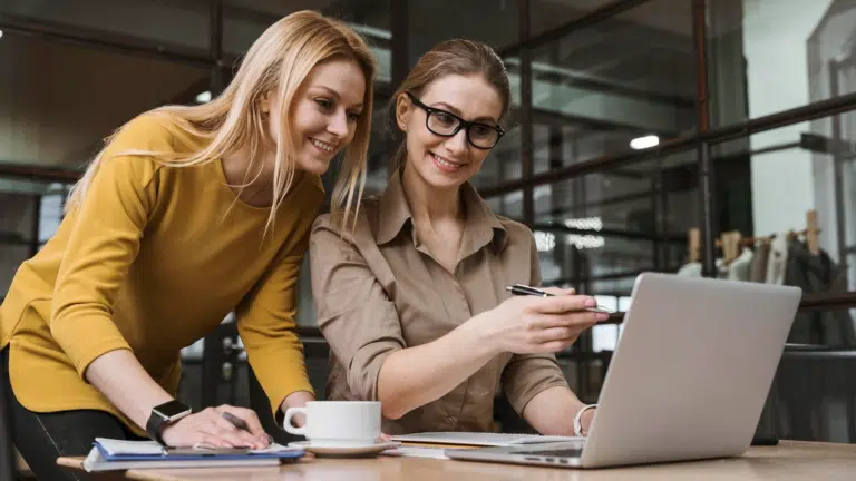 Two female works collaborate over a laptop in an office environment. They both smile.