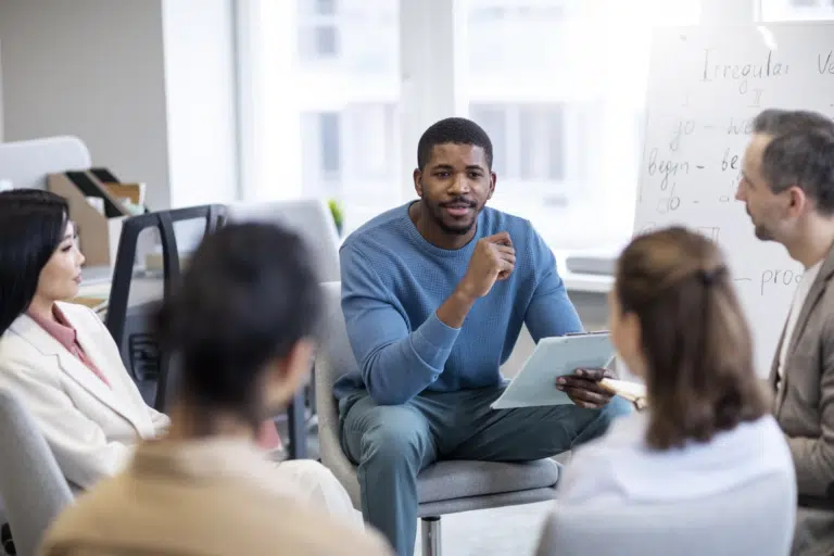 A black, male in a blue jumper refers to a clipboard in his hands whilst talking to 4 other people in a circle. Behind him is a white board that indicates 'Irregular Verbs'