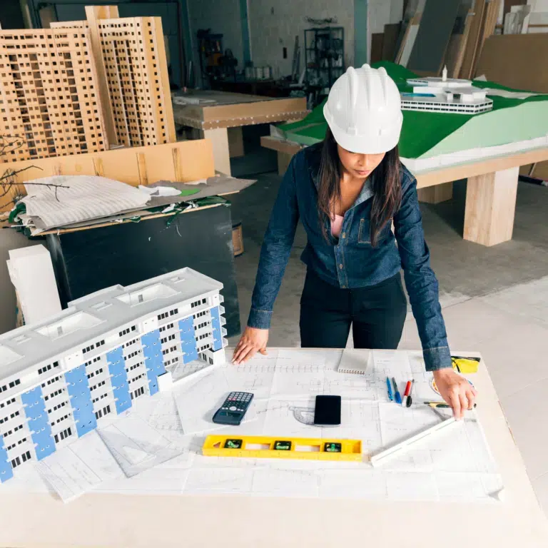 A female construction worker wearing a white hard hat and a denim jacket looks over construction plans in a room of models.