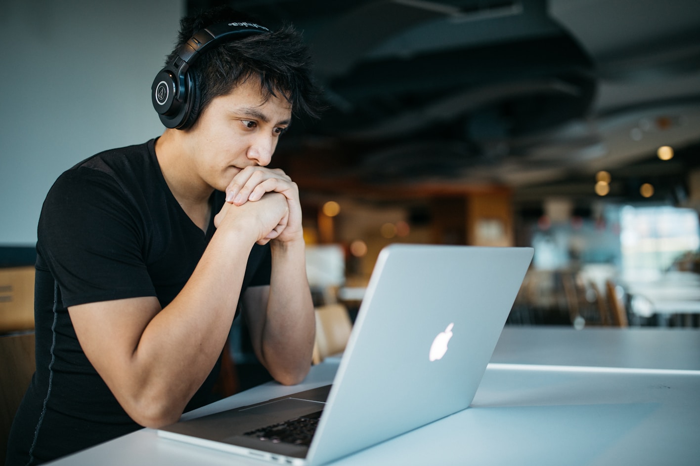 An Asian male rests his chin on his hands as he looks down at a laptop. He wears over ear headphones and is concentrating.