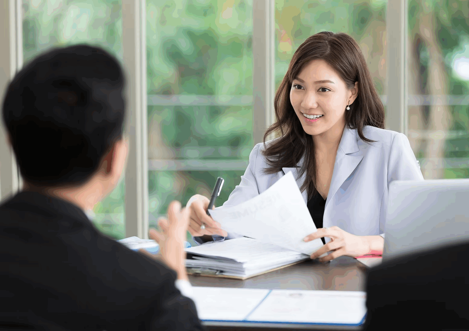 An Asian female in a lilac suit jacket interviews a man opposite her. She smiles, has long brown hair and holds a pen and a CV in her hand.