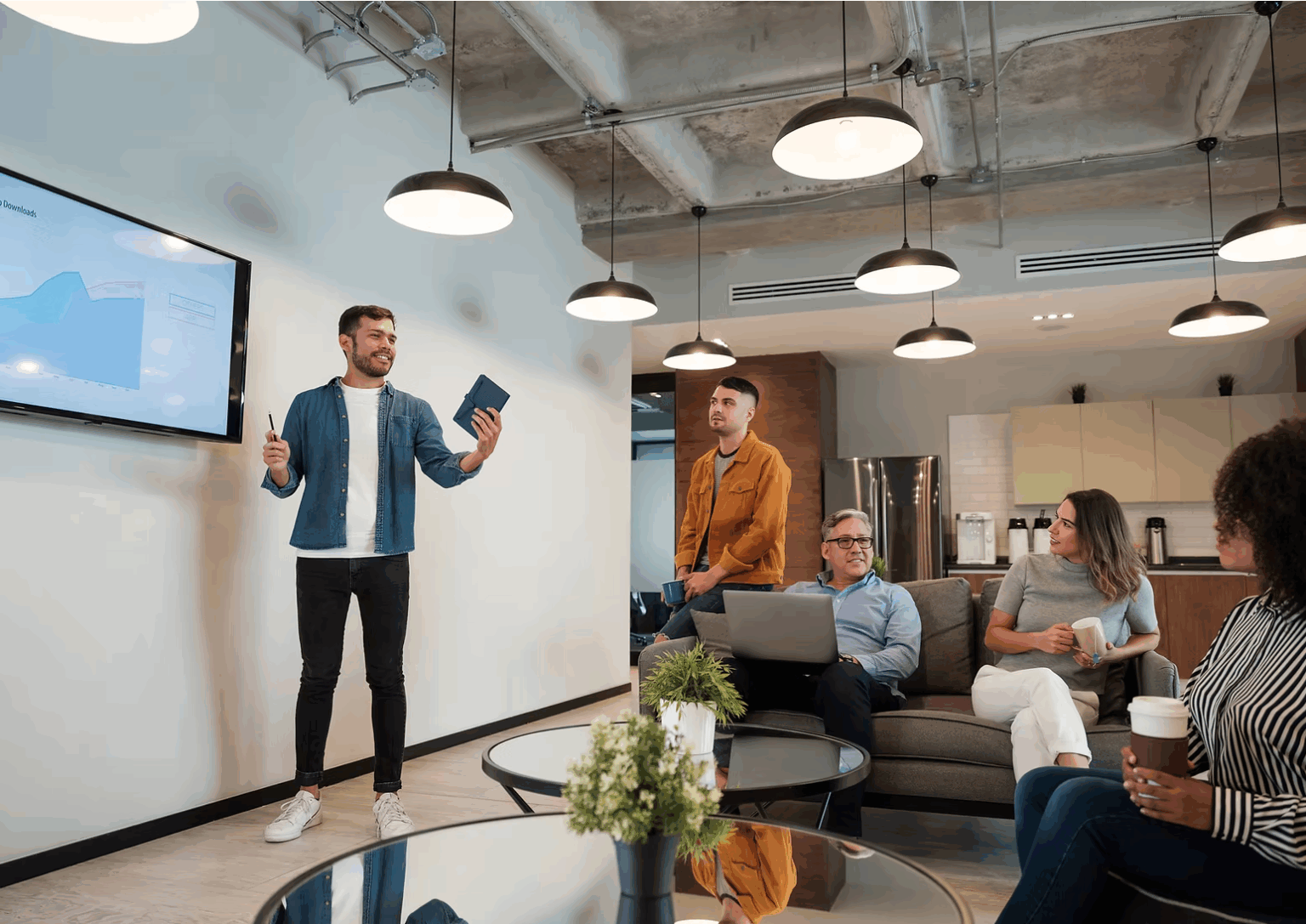 A white male address his colleagues in a meeting in an open plan office with sofas and glass coffee tables. He is animated and smiling.