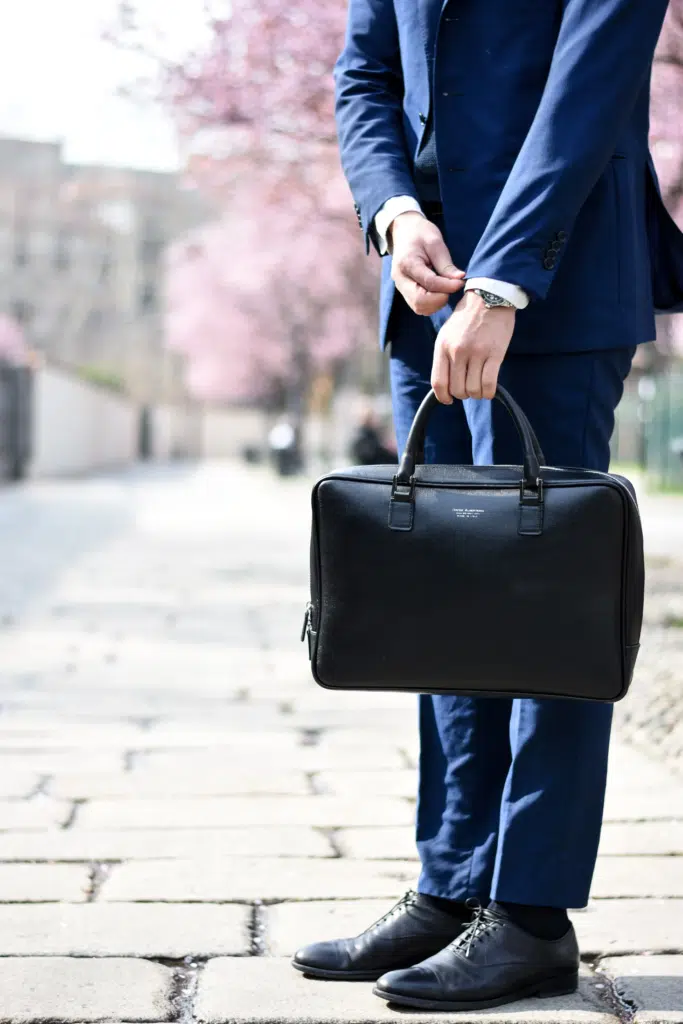 A man stands on a blossom-tree lined street holding a brief case. He fiddles with the cuff on his suit.