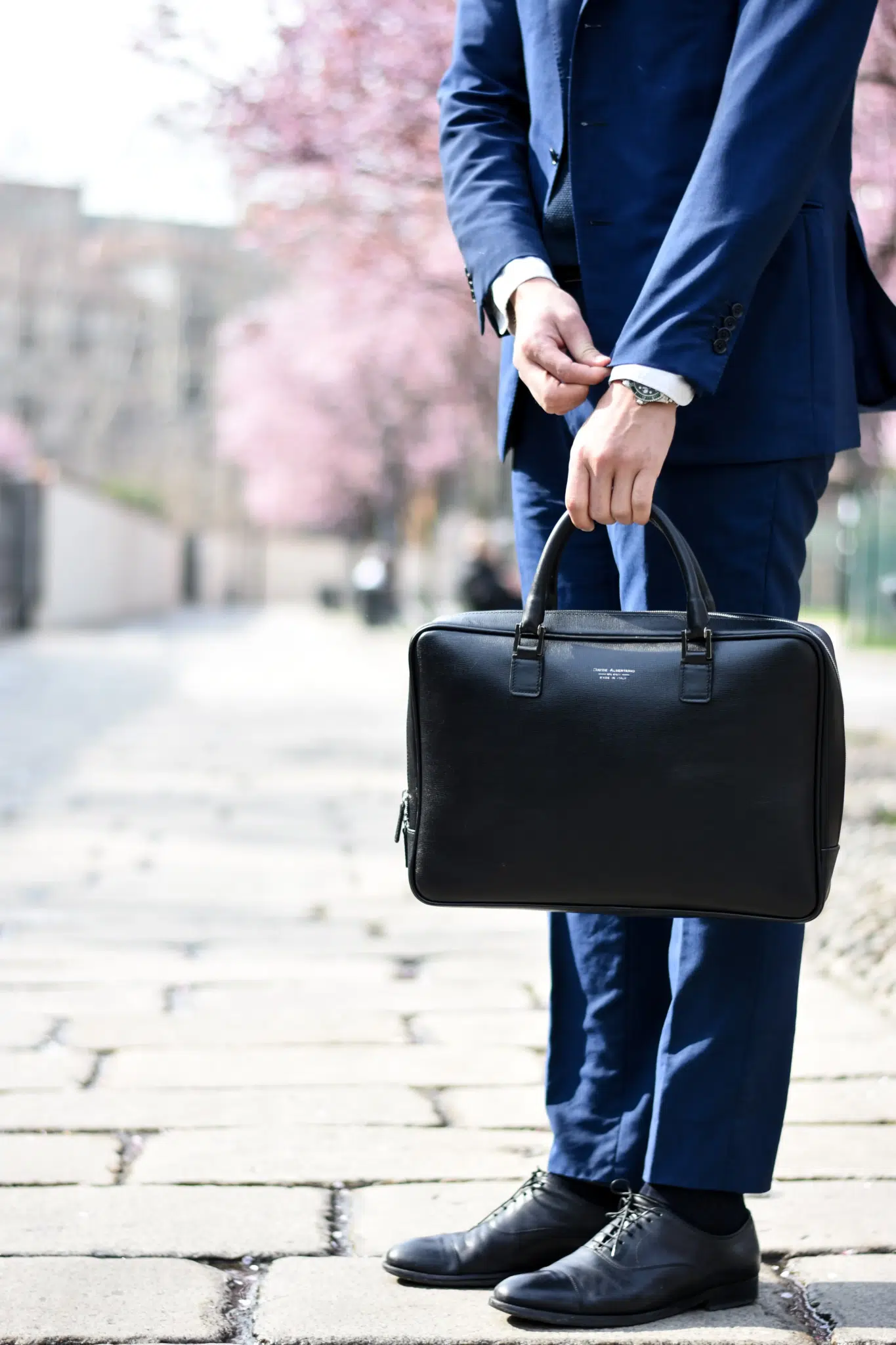 A man stands on a blossom-tree lined street holding a brief case. He fiddles with the cuff on his suit.