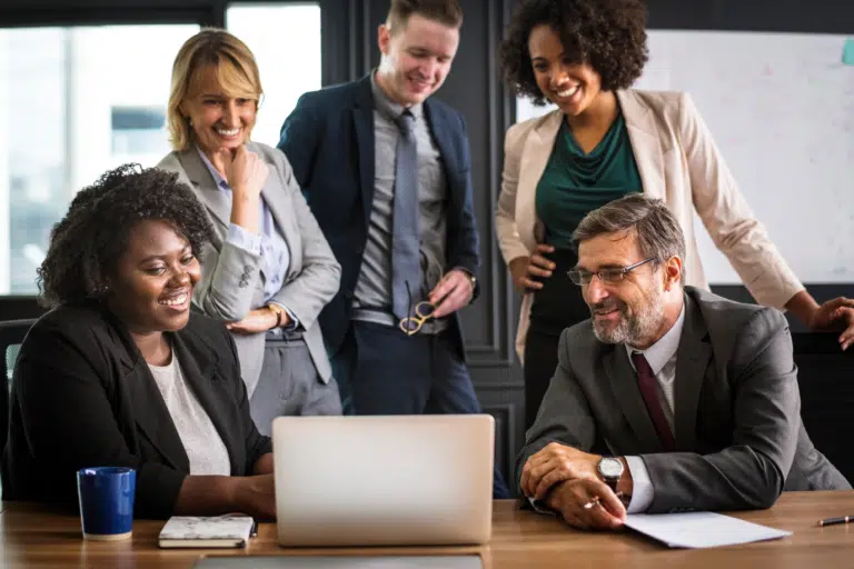 A diverse group of workers look happy and pleased around a laptop in an office. They all wear formal office wear.