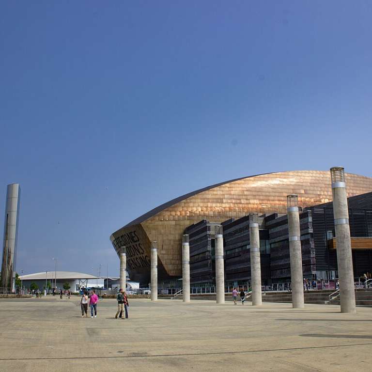 A large, bronze, curved (almost domed) industrial building. It is a bright day - no clouds are in the blue sky. People wander in front of the impressive building.