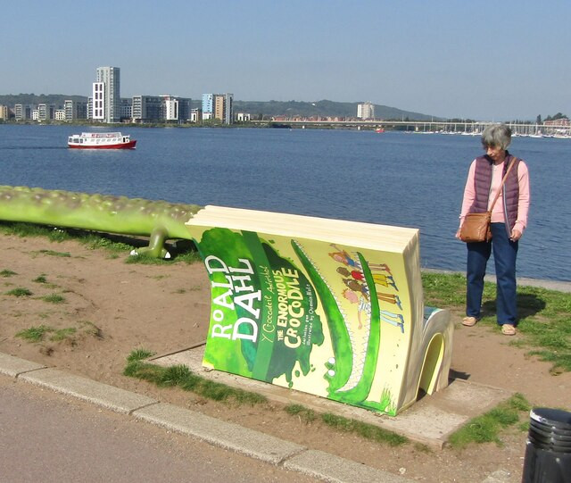 An older female in a pink top and gilet looks at a bench that's been made in the shape of a Roald Dahl book. The bench looks over a large body of water with boats in the background.