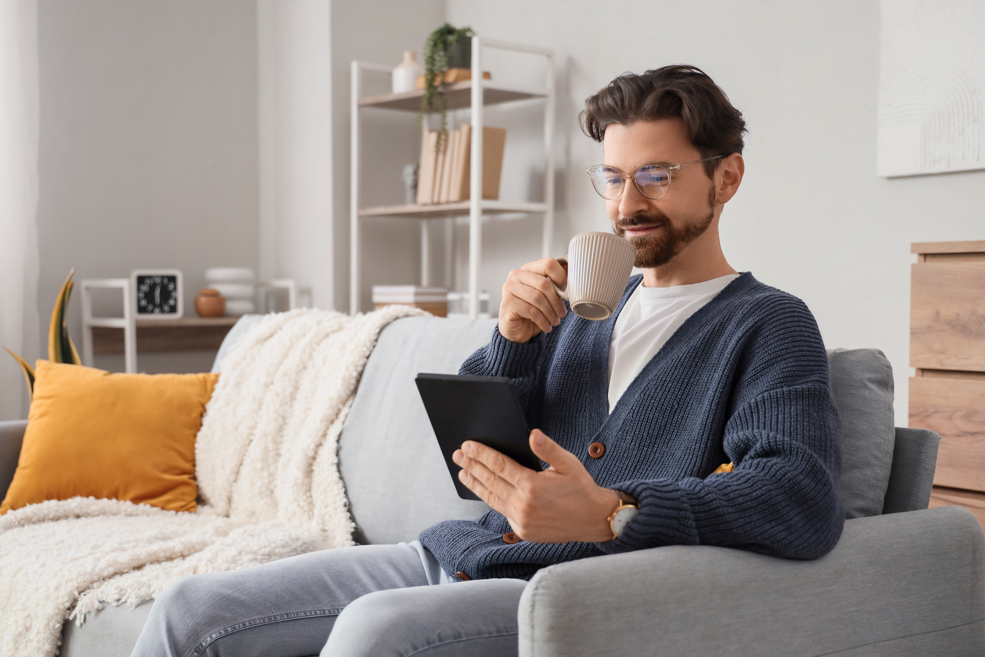 A young ma sits and reads a Kindle sized device and drinking a cup of coffee. He's sat on a sofa in a modern looking, white and bright living room.