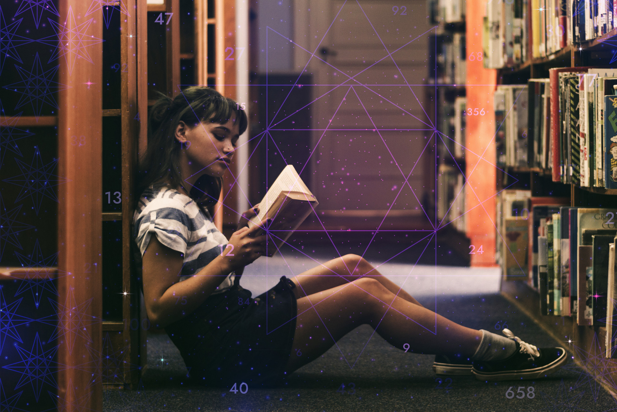 A young, female student sits and reads a book in a library on the floor leant against a book shelf. She wears a black denim skirt, white stripey top and black sneakers with grey socks.
