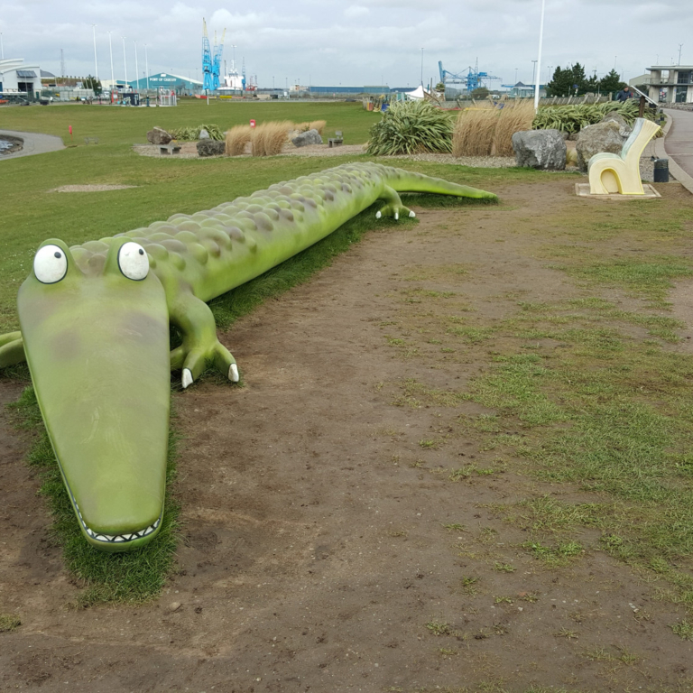 A large plastic crocodile sits next to a lagoon on a green. It is a cloudy day.