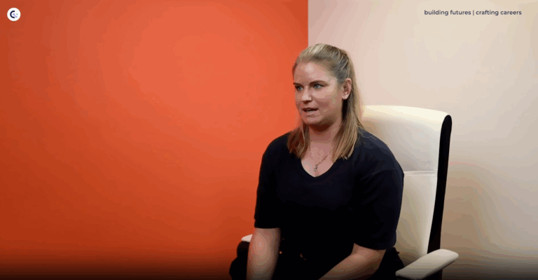 Charlotte Farmery - a white female with shoulder length blonde hair - sits in a white office chair talking against an orange background